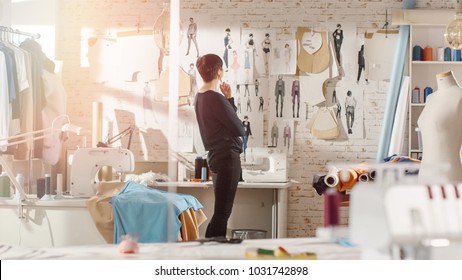 Female fashion, designer,  Looking at Drawings and Sketches that are Pinned to the Wall Behind Her Desk. Studio is Sunny. Personal Computer, Colorful Fabrics, Sewing Items are Visible.