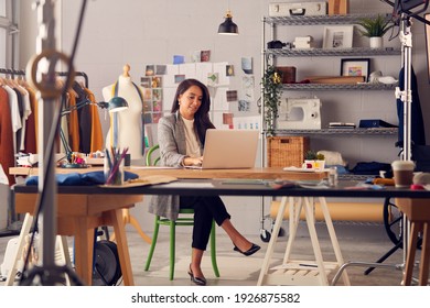 Female Fashion Designer In Studio Working On Sketches Or Documents At Desk With Laptop