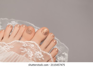 Female Feet With Beige Nail Design. Glitter Beige Nail Polish Pedicure With Black Dots Nail Art. Woman Feet With Perfect Pedicure And White Lace On Gray Background. Copy Space.