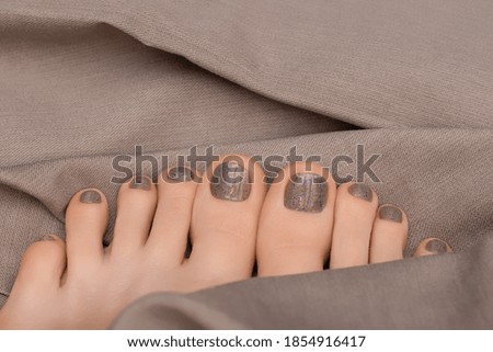 Female feet with beige nail polish. Gray pedicure on female feet. Woman legs with gray nail design on beige fabric