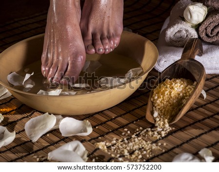 Female feet with drops of water, spa bowl, towels, flowers, candles, sea salt and bottle with essential oil. Foot spa concept.