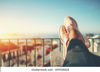 Female feet with glossy pale pink shoes relaxing on wooden table and touristic european city near sea with multiple houses in blurred background with copy space for advertising, logo or your texts