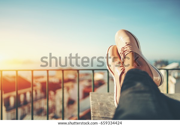Female feet with glossy pale pink shoes relaxing on\
wooden table and touristic european city near sea with multiple\
houses in blurred background with copy space for advertising, logo\
or your texts