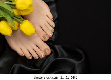 Female feet with golden nail polish pedicure. Woman legs with glitter golden nail design and yellow tulip flowers on black fabric background. Copy space.