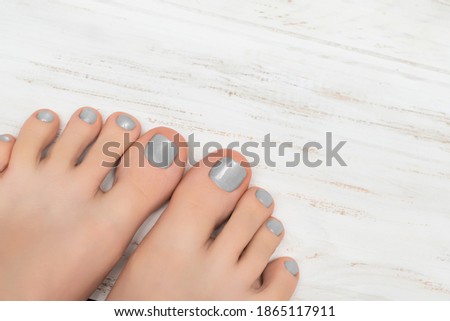 Female feet with gray nail polish. Glitter pedicure on female feet. Woman with gray pedicure on white wooden desk.