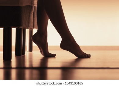 Female Feet On Hardwood Floor. Young Woman Waking Up And Getting Up From Bed In The Morning. Silhouette Of Legs And Body In Bedroom.