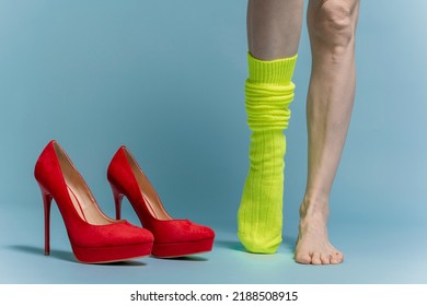 Female Feet With One Green  Sock Standing Next To Red High Hills. Isolated On Blue Background