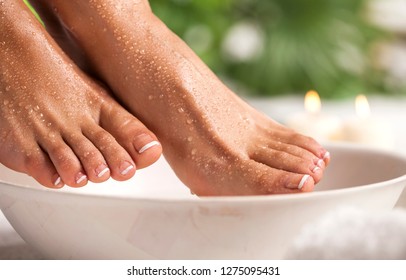 Female feet with spa bowl, towel, soap and flowers on white floor and tropical green leaves background