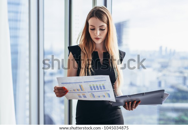 Female financial analyst\
holding papers studying documents standing against window with city\
view
