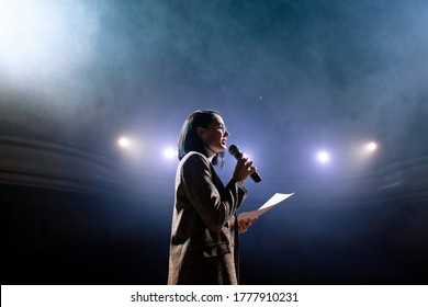 The female financial coach emotional gesturing talks from the stage with spectators at forum. Too many anonymous persons workers and students seat in large auditorium and watch workshop background - Shutterstock ID 1777910231