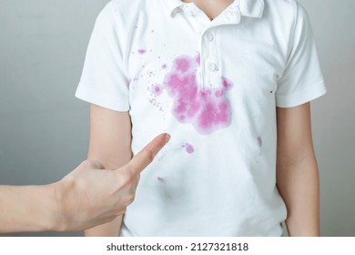 Female Finger Points To A Dirty Stain Spilled Juice On A White Children's T-shirt. The Concept Of Cleaning Stains On Clothes. 