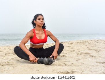 Female Fitness Athlete Stretches On The Beach With Arms Pressed On Her Legs To Stretch Groin