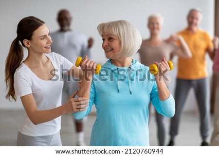 Female fitness coach helping active senior woman while exercising with dumbbells, sporty multiracial group of elderly people having fitness class at nursing home, training with instructor