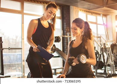 Female fitness instructor showing exercise progress on clipboard to young athletic woman at gym.
