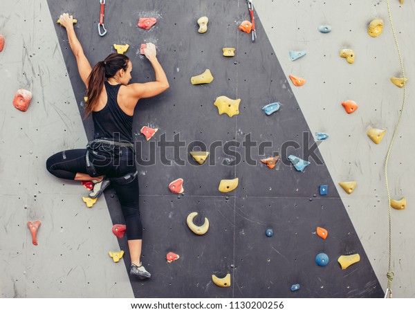 Female fitness professional climber training at\
bouldering gym. Muscular woman with athletic body dressed in black,\
climbing on artificial colourful rock wall. Active lifestyle and\
bouldering concept.