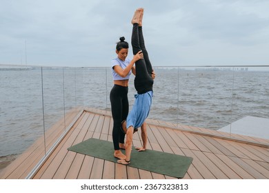 Female fitness trainer helps young man to do handstand at embankment on exercise at with view on ocean by holding guy's legs. Training outside, healthy lifestyle. Couple training together. - Powered by Shutterstock