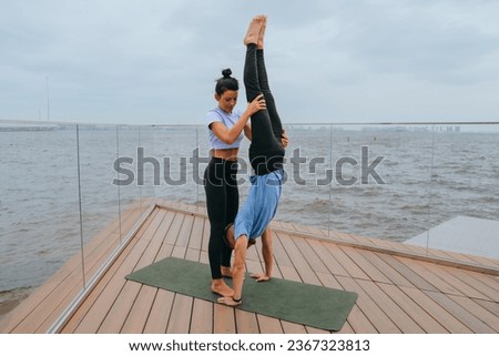 Female fitness trainer helps young man to do handstand at embankment on exercise at with view on ocean by holding guy's legs. Training outside, healthy lifestyle. Couple training together.