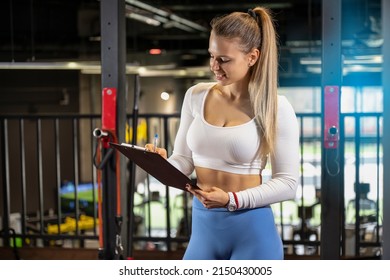 Female Fitness Trainer Holding Clipboard And Writing Sports Report For A Client In The Gym