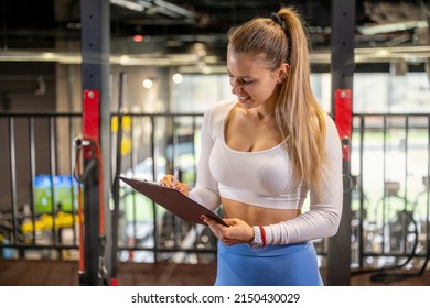 Female Fitness Trainer Holding Clipboard And Writing Sports Report For A Client In The Gym