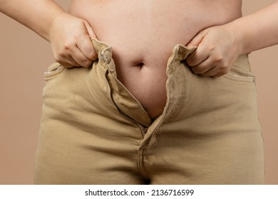 Female flabby big tummy preventing her from putting jeans on on beige background. Visceral fat. Body positive. Sudden weight gain. Tight little clothes. Need for wardrobe change.