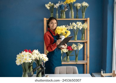 A female florist holds a bouquet and makes notes on a clipboard while standing in her flower shop. Small business and floristry concept - Powered by Shutterstock