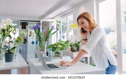 Female Florist Standing At Her Flower Shop Counter Using Mobile Phone And Laptop To Take Orders For Her Store. Woman Flower Shop Owner Taking Online Orders.