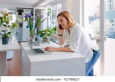 Female florist talking on mobile phone while using laptop in flower shop. Happy young woman taking order in plant nursery. - Powered by Shutterstock