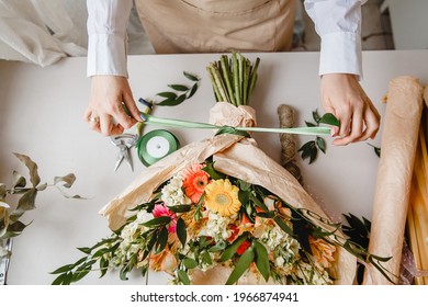 a female florist ties a ribbon bow on a bouquet of flowers wrapped in craft paper on the desktop. Top view. High quality photo - Shutterstock ID 1966874941