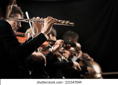 Female Flutist Close-up With Orchestra Performing On Background.