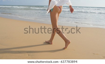 Female foot stepping on the sand with sea waves background. Beautiful woman in swimsuit and shirt walking on sea beach barefoot. Young girl going on the ocean shore. Summer vacation concept. Close up.