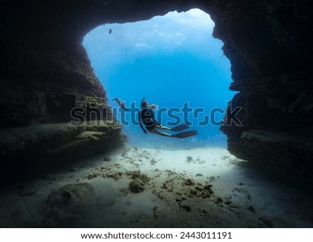 A female free diver observes a Pacific green sea turtle framed by an underwater cave opening in the clear blue waters of Hawaii. Model release provided.