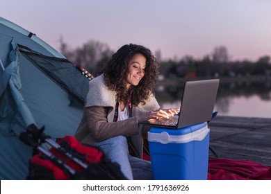 Female Freelancer Camping By The Lake, Sitting On The Tent Entrance And Working On A Laptop Computer;  Digital Nomad Lifestyle