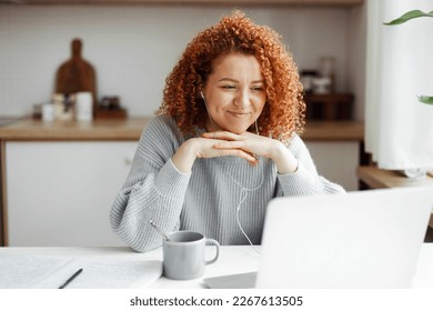 Female freelancer having video conference with her employer discussing new project details, listening to him attentively in headphones smiling at web camera, putting folded hands under chin