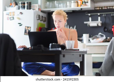 Female freelancer in her casual home clothing working remotly from her dining table in the morning. Home kitchen in the background.