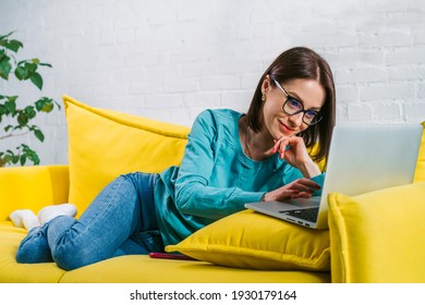 Female freelancer working from home using her latop sitting on comfortable yellow sofa. Distant work or education, self-isolation concept.