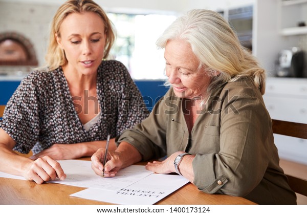 Female Friend Helping Senior Woman To Complete Last\
Will And Testament At\
Home