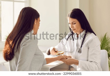 Female friendly doctor examining hand skin of young woman patient. Professional dermatologist using magnifying glass checking skin condition in clinic. Dermatology, skin health care concept.