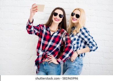 Female friends in checkered shirts making selfie portrait with phone on the white wall background