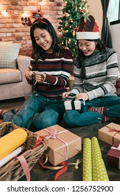 Female Friends Having Fun Doing Art Work Wrapping Gifts. Young Girl Using Scissors Cutting Green Ribbon Handmade Presents. Besties Prepare Celebration Christmas Eve New Year Beside Xmas Tree.