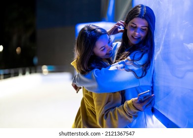 Female Friends Hugging Next To A Blue Neon Wall At Night Holding Their Mobile Phones And Laughing