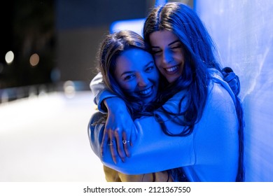 Female Friends Hugging Next To A Blue Neon Wall At Night Holding Their Mobile Phones And Laughing