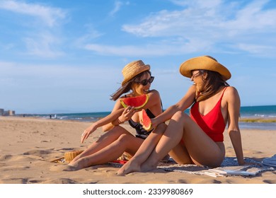 Female friends on vacation at the beach in summer eating a watermelon with the sea in the background, sitting on the sand - Powered by Shutterstock