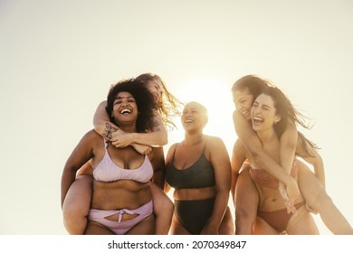 Female Friends Piggybacking Each Other At The Beach. Group Of Happy Young Women Smiling Cheerfully While Standing In The Summer Sun. Best Friends Having Fun And Enjoying Their Vacation Together.