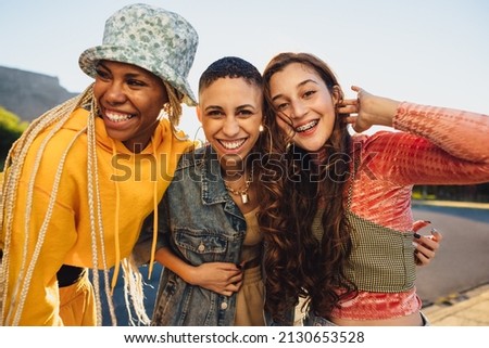 Female friends smiling cheerfully while embracing each other. Female youngsters having fun while standing together outdoors. Group of generation z friends making happy memories together.