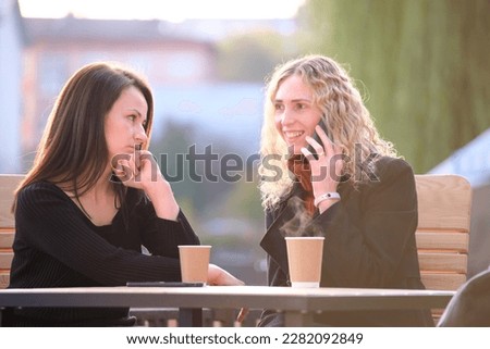Female friends spending time at street cafe outdoors while one girl is talking happily on mobile phone and another waiting frustrated at offensive behaviour of her mate. Friendship problems concept