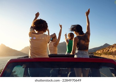 Female Friends Standing Up Through Sun Roof Car And Dancing On Road Trip Through Countryside - Powered by Shutterstock