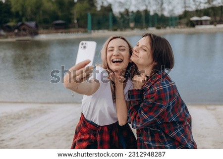 Female friends take selfies in nature on shore of lake, using a mobile app for social media. The girls are having a fun summer vacation in the heart of nature, capturing videos on their smartphones.