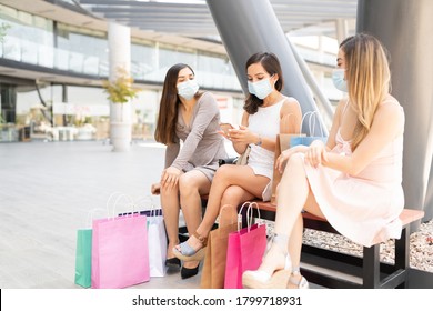 Female Friends Wearing Face Masks While Sitting On Bench At Shopping Center