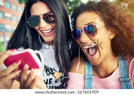 Female friends wearing sun glasses and smile widely as they take a photo with their cell phone