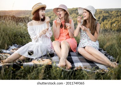 Female Friendship. Picnic Time. Three Attractive Girls Holding Escargot Snails In Hands And Eating, Against The Background Of Green Hills. Blanket With Food Spread On It.